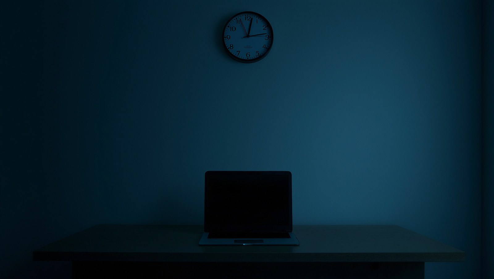 A dark empty office at night, a laptop with a black screen on the desk and a wall clock above it